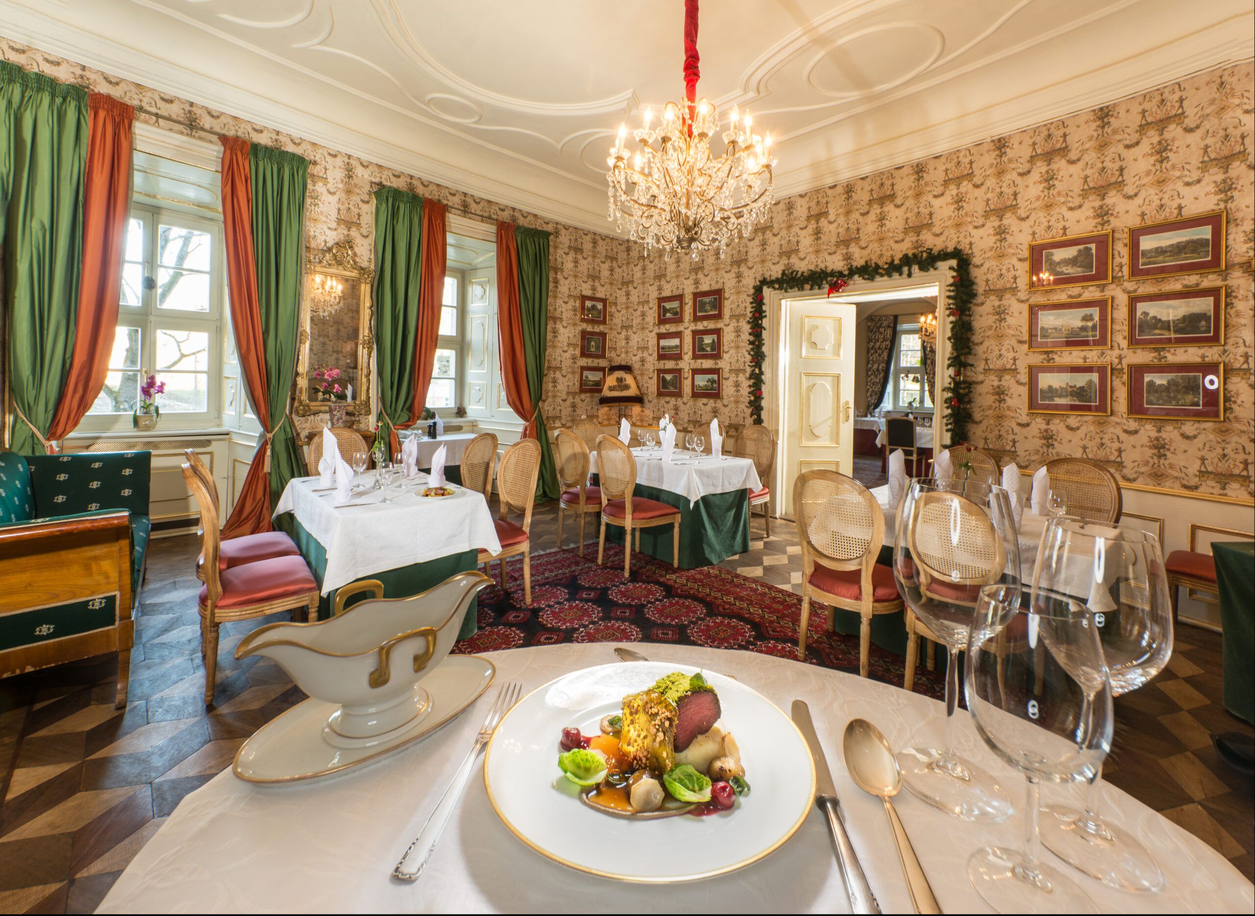 Wnętrze restauracji w stylu historyzującym, Na oknach duże, kotarowe zasłony. Stoły zastawione, przyszykowane na uroczystość. Na ścianach wzorzyste tapety oraz dużo zdjęć w ramach. Drewniany parkiet, przykryty prostokątnym dywanem we wzór geometryczny. Na pierwszym planie talerz z wykwintnym posiłkiem.