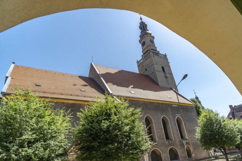 Widok na parafię w Kamiennej Górze, szara elewacja, z czerwonawym dachem, na pierwszym planie kilka drzew. W tle niebieskie niebo.