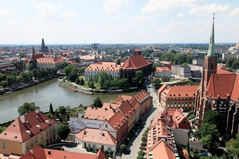 Panorama miasta z zabytkowymi budynkami pokrytymi czerwonymi dachówkami, znajduje się wyspa i przepływająca rzeka Odra