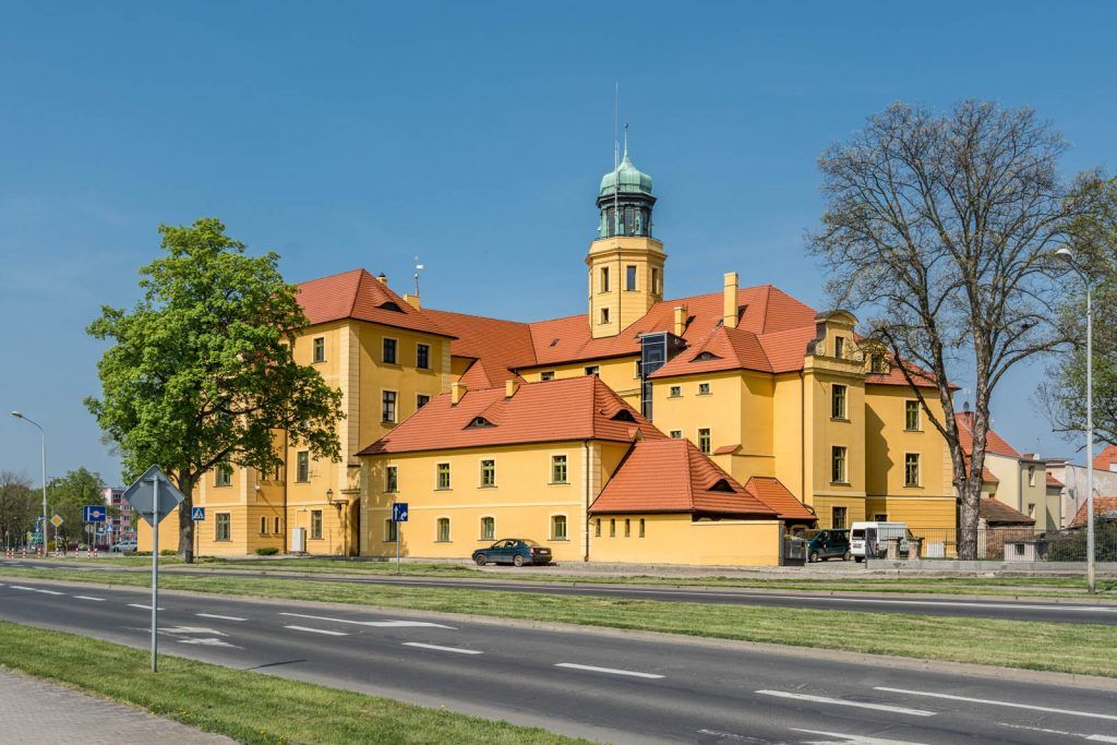Ceglana, czerwonawo-brązowa budowla z jasnymi detalami wokół okien i bram, wyróżniająca się wieżą w kolorze czerwonej cegły.