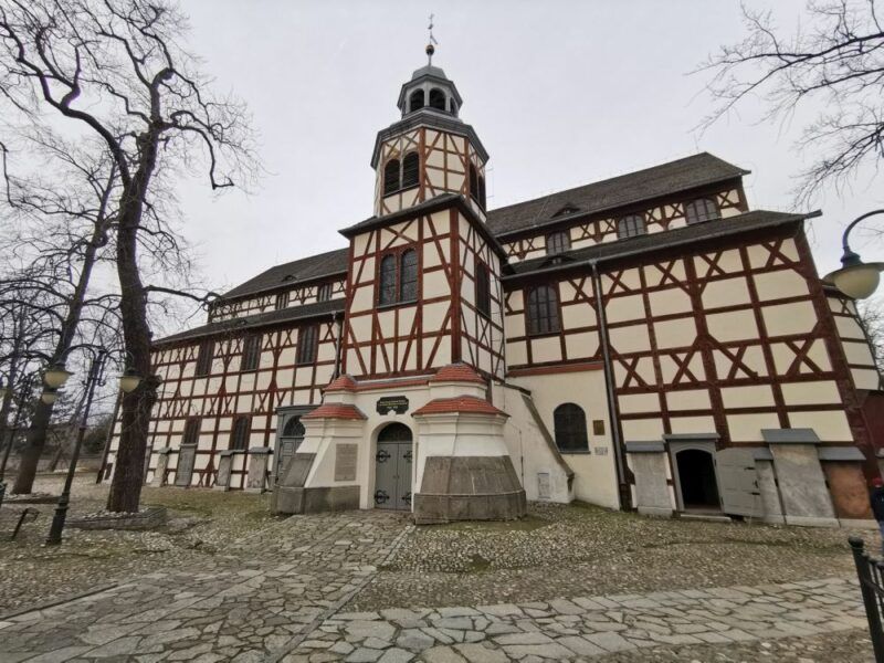 Na zdjęciu widoczny jest Kościół Pokoju w Jaworze, charakterystyczna budowla zbudowana z drewna, wpisana na listę UNESCO. Fasada kościoła wyróżnia się biało-brązowym układem drewnianych belek oraz centralną wieżą z barokowym hełmem. Otaczają go stare, bezlistne drzewa oraz brukowana ścieżka prowadząca do wejścia.