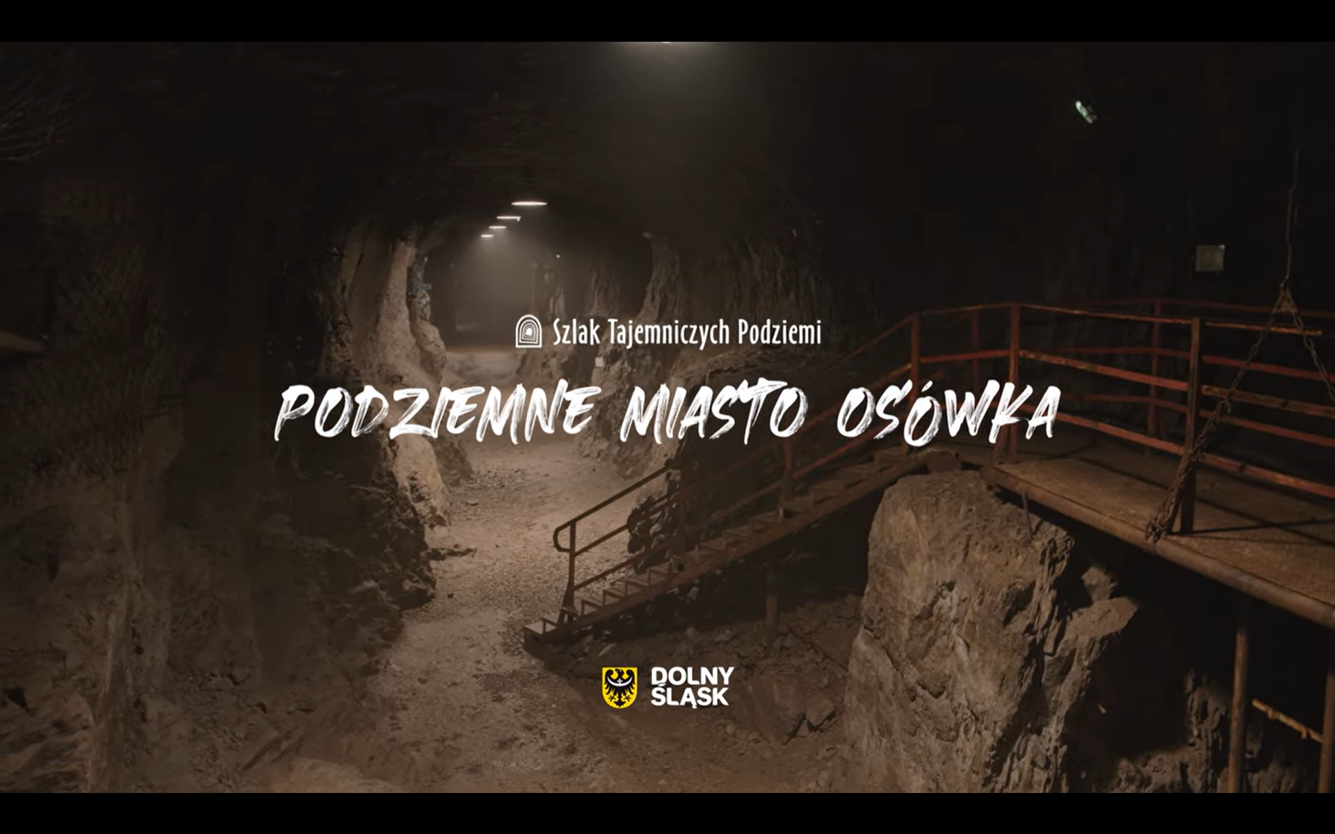 wysoki oraz długi wydrążony korytarz, lekko oświetlony. po prawej stronie znajdują się ciemno czerwone schody które prowadzą do wyżej położonego tunelu