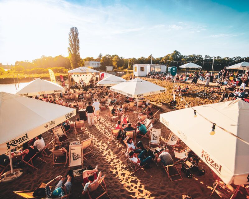 Zdjęcie wykonane przy słonecznej, letniej pogodzie, przedstawia bar na plaży z gośćmi siedzącymi na leżakach.