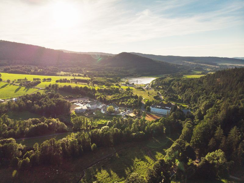 Zdjęcie lotnicze przedstawiające zielone tereny leśne, na tle niewielkich szczytów gór oraz skryte pomiędzy nimi małe zabudowania. W oddali zbiornik wodny.