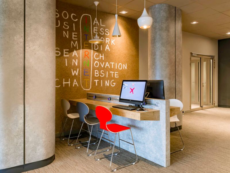 Lobby hotelowe w nowoczesnym modernistycznym stylu, ze stołem barowym z komputerami oraz nowoczesnymi krzesłami. Na ścianie modernistyczna fototapeta.