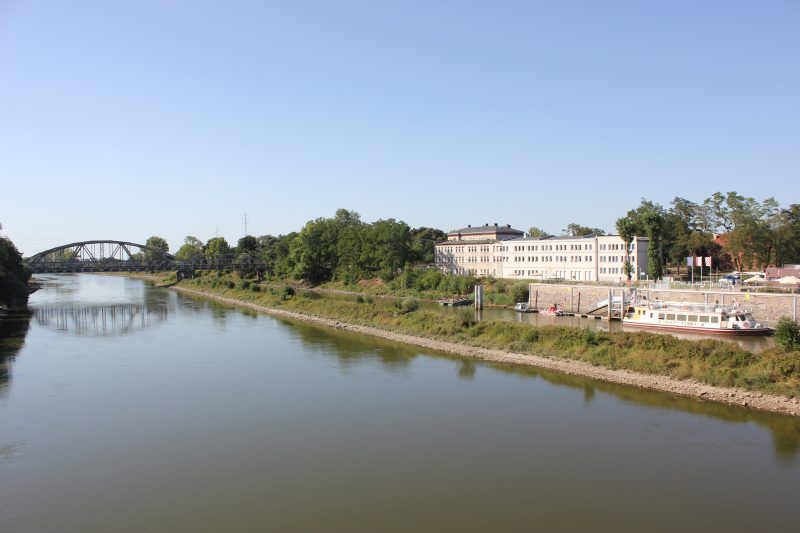 widok z góry na szeroką rzekę, po drugiej stronie rzeki znajduję się biały podłużny budynek w oddali widać brązowy most