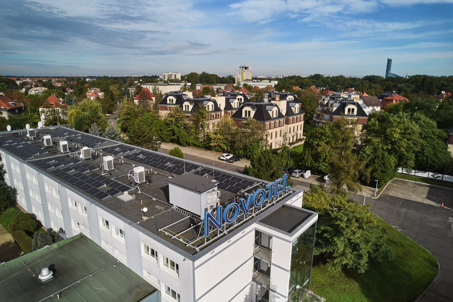 Widok z góry na budynek oznaczony niebieskim napisem „Novotel”, umieszczonym na dachu. W tle rozciągają się drzewa, osiedla i samotna wieża. Niebo jest lekko zachmurzone, nadając scenie spokojny nastrój. Hotel znajduje się pośrodku zielonego, miejskiego krajobrazu,
