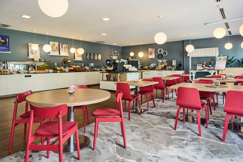 Zdjęcie przedstawia wnętrze nowoczesnej restauracji. W pomieszczeniu dominują czerwone krzesła, drewniane stoły i wiszące lampy sufitowe. W głębi znajduje się bar z butelkami i szklankami. Wystrój tworzy elegancki i przyjazny klimat.