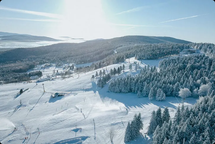 Rozległy ośnieżony stok narciarski biegnie przez łagodne, porośnięte świerkami wzgórza, z trasami, wyciągami i pojedynczymi zabudowaniami w dolinie.