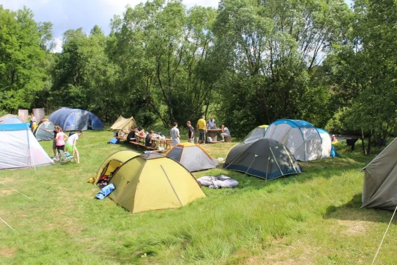 Kolorowe namioty stoją na trawiastej polanie pośród drzew, gdzie turyści odpoczywają i przygotowują obozowisko na niewielkim kempingu.