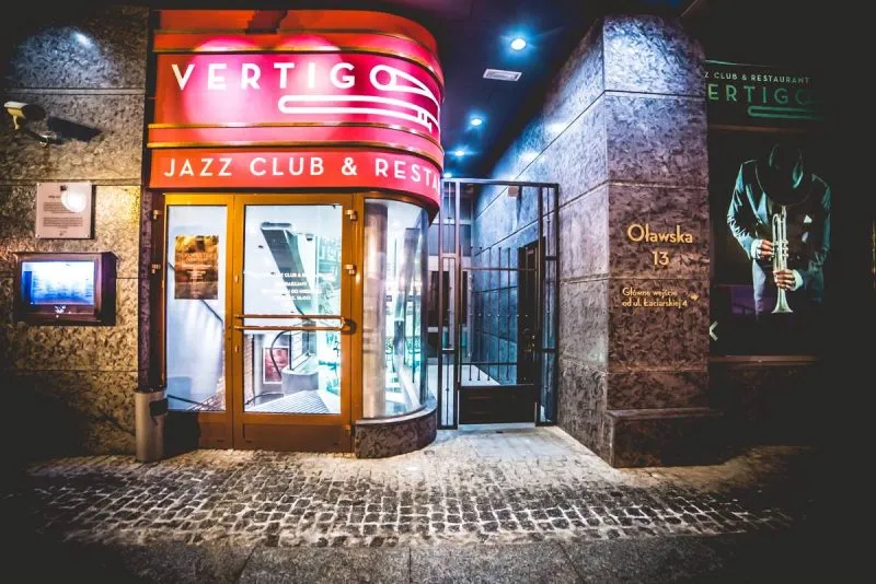 Nocne wejście do Vertigo Jazz Club and Restaurant we Wrocławiu rozświetlają czerwone i zielone neony nad oszklonymi drzwiami w kamiennej fasadzie.
