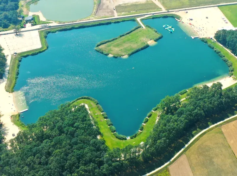 Widok z lotu ptaka ukazuje turkusowe jezioro otoczone lasem i polami, z piaszczystymi plażami, trawiastą wyspą oraz pływającym torem przeszkód przy brzegu.
