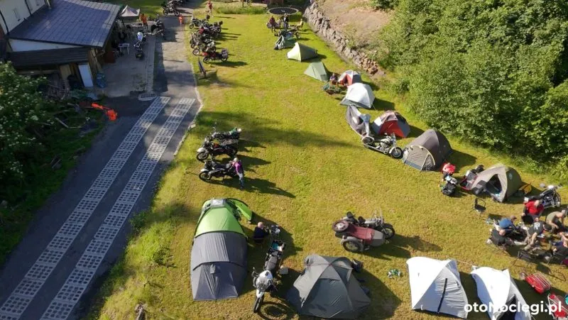Namioty i motocykle turystyczne stoją na trawiastej polanie przy gospodarstwie agroturystycznym, ustawione w dwóch rzędach wzdłuż brukowanej alejki.