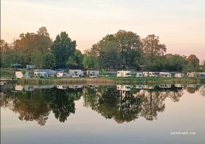 Kampery i przyczepy stoją wokół spokojnego stawu na zielonym polu caravaningowym, a w tafli wody odbijają się rozłożyste drzewa i wieczorne niebo.