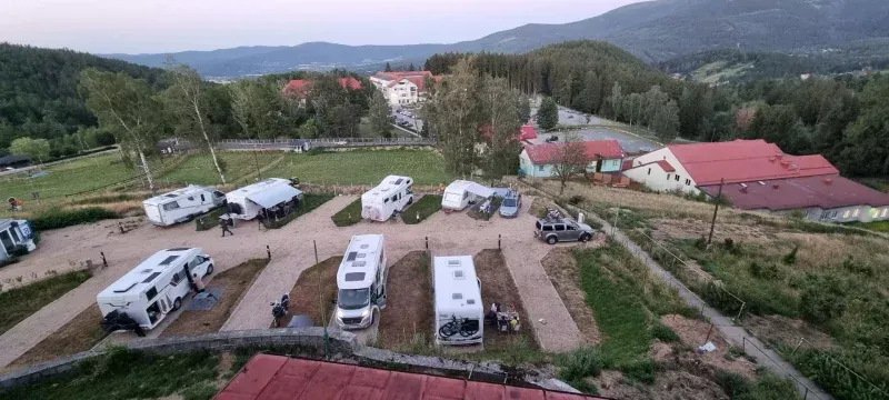 Górskie pole caravaningowe widziane z góry, z rzędami kamperów i przyczep rozstawionych na tarasach pośród zabudowań, drzew oraz zalesionych wzgórz.