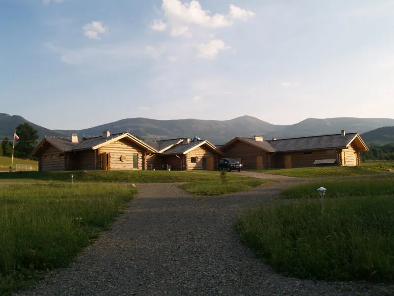 A row of wooden cabins at the Camp66 campsite stands on a wide meadow at the foot of gentle, forested mountains under a bright, partly cloudy sky.