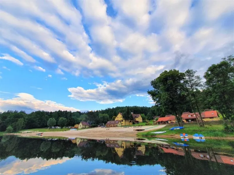 Spokojny staw przy piaszczystej plaży odbija błękitne niebo, a na zielonym brzegu stoją niewielkie domki, leżaki i sprzęt wodny dla gości.