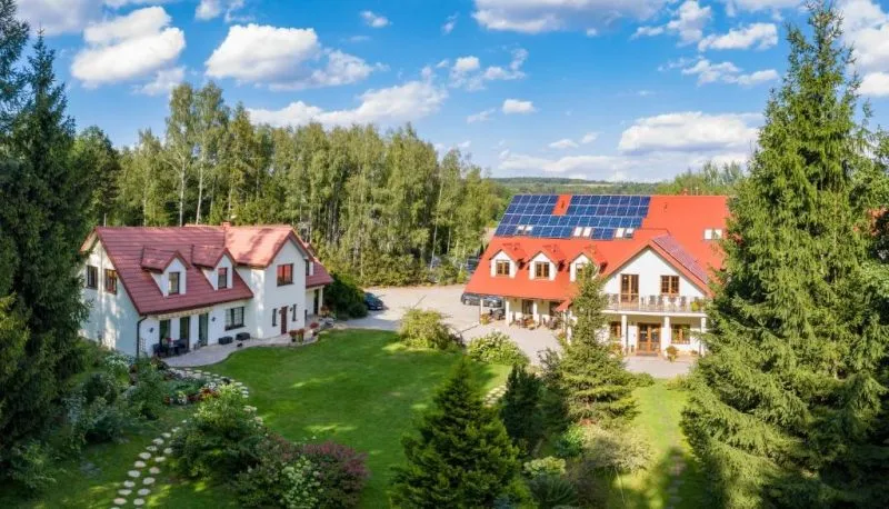 Dwa jasne pensjonaty z czerwonymi dachami, panelami słonecznymi i zielonym ogrodem stoją na skraju lasu, tworząc spokojną bazę dla caravaningu.