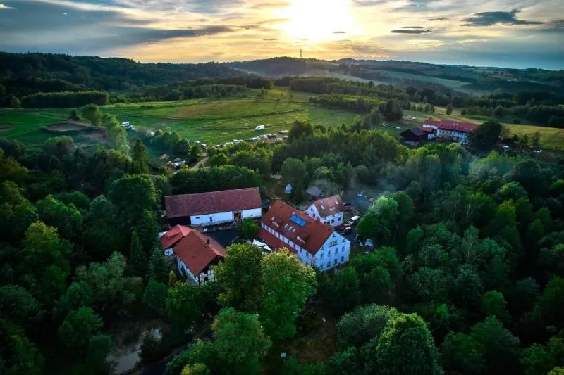 Widok z drona o zachodzie słońca ukazuje wiejski kompleks zabudowań pośród pól, łąk i zalesionych wzgórz, z wijącymi się drogami w dolinie.