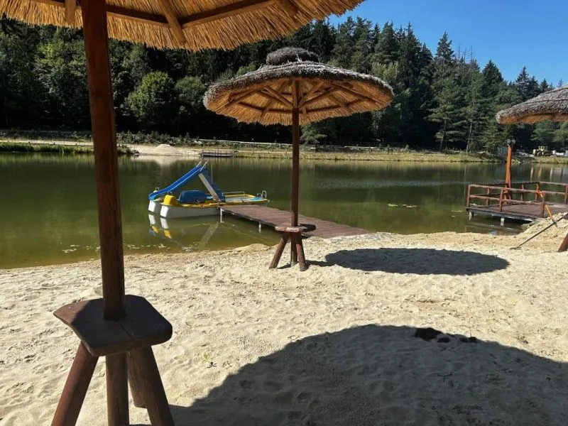 Piaszczysta plaża nad leśnym stawem ma słomiane parasole, drewniane pomosty i sprzęt wodny, a spokojną taflę otaczają wysokie drzewa.