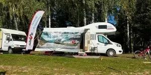Dwa białe kampery promocyjne stoją na zielonej leśnej polanie przy wysokiej fladze, a obok przygotowano rower i niewielkie stanowisko turystyczne.