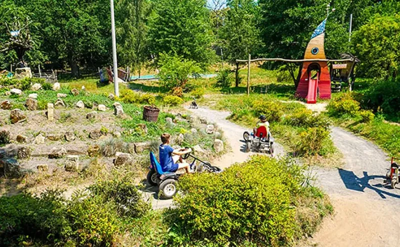 Children ride small off-road vehicles along the paths of a green camping park, passing rock gardens, bushes, and a wooden house.