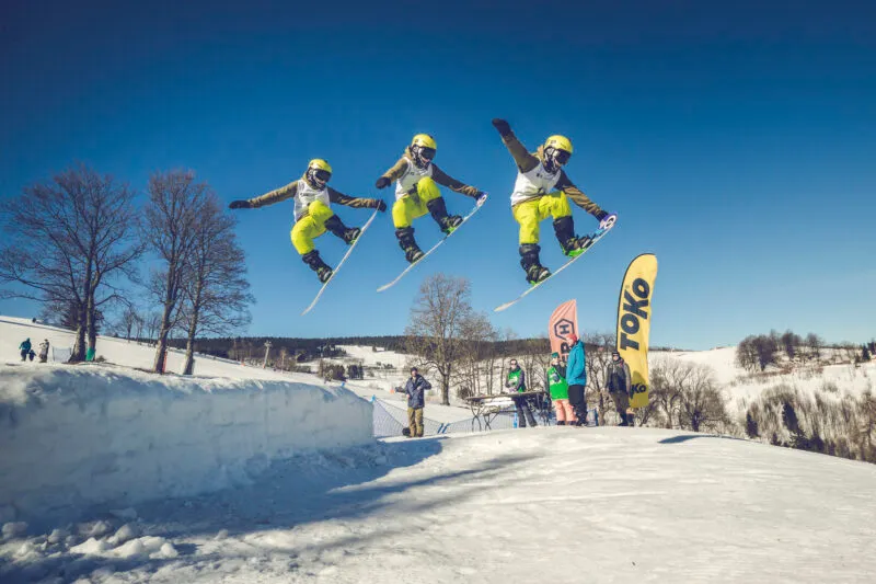 Snowboardzista wykonuje wysoki skok nad ośnieżonym stokiem na Ziemi Kłodzkiej, pokazany w trzech kolejnych fazach ewolucji na tle błękitnego nieba.