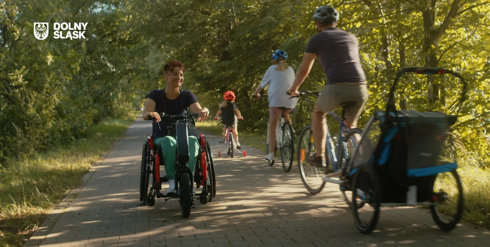 Osoby jadą szeroką parkową aleją korzystają z rowerów, handbike’ów i przyczepek, pokazując dostępną turystykę dla rodzin i osób z niepełnosprawnościami.