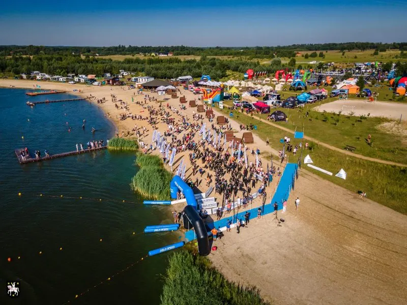 Widok z drona na zatłoczoną plażę nad jeziorem, pole namiotowe i kolorowe strefy rekreacyjne z pomostami oraz sprzętem wodnym.