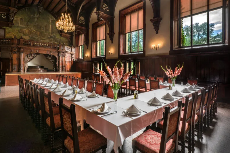 Restauracyjna sala Zamku Kliczków ma długi elegancko nakryty stół, drewniane boazerie, wysokie okna, rzeźbiony kominek i kryształowy żyrandol.