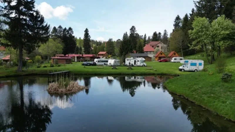 Przyczepy kempingowe i namioty rozstawione wokół spokojnego stawu, w którego tafli odbijają się drzewa, trawiasty brzeg oraz zabudowania ośrodka caravaningowego.