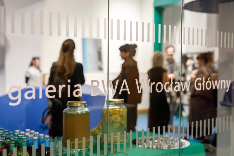 Przez witrynę galerii BWA Wrocław Główny widać kolorowe butelki ustawione na ekspozycji oraz rozmawiających gości w jasnym wnętrzu Palmiarni.