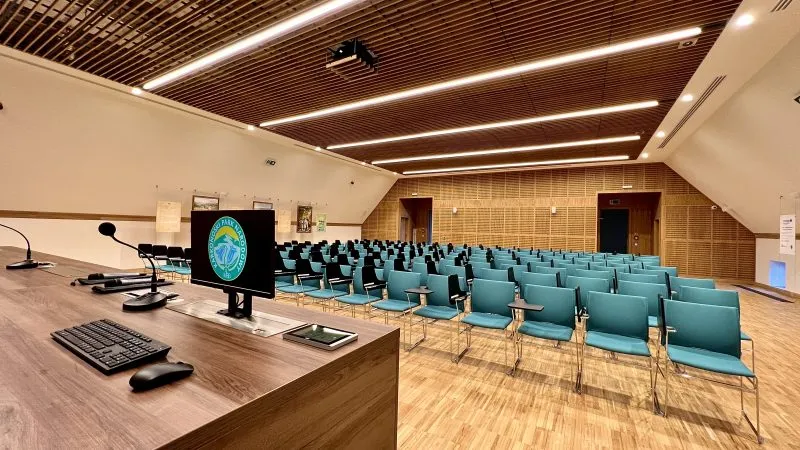 Nowoczesna sala konferencyjna z widokiem z perspektywy mównicy. Na pierwszym planie biurko z mikrofonami, klawiaturą i monitorem z logo Karkonoskiego Parku Narodowego. Przed biurkiem ustawione rzędy turkusowych krzeseł z składanymi blatami. Wnętrze wykończone zostało drewnem.