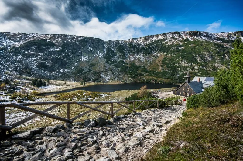 Zdjęcie wykonane ze szlaku pieszego przedstawia panoramę Karkonoszy, staw oraz drewniany budynek schroniska