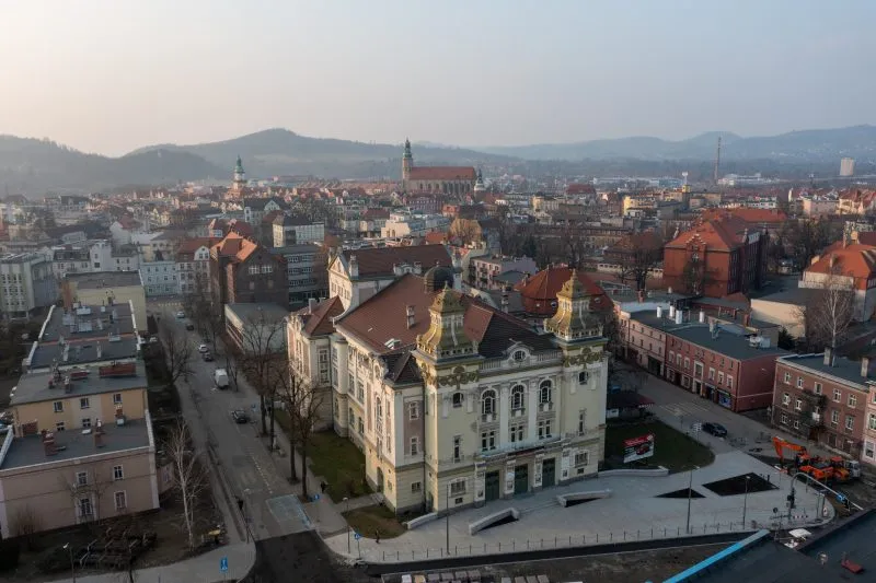 Panorama centrum Jeleniej Góry widziana z góry, z gmachem teatru, zabytkowymi kamienicami, wieżami kościołów i górskimi pasmami na horyzoncie.