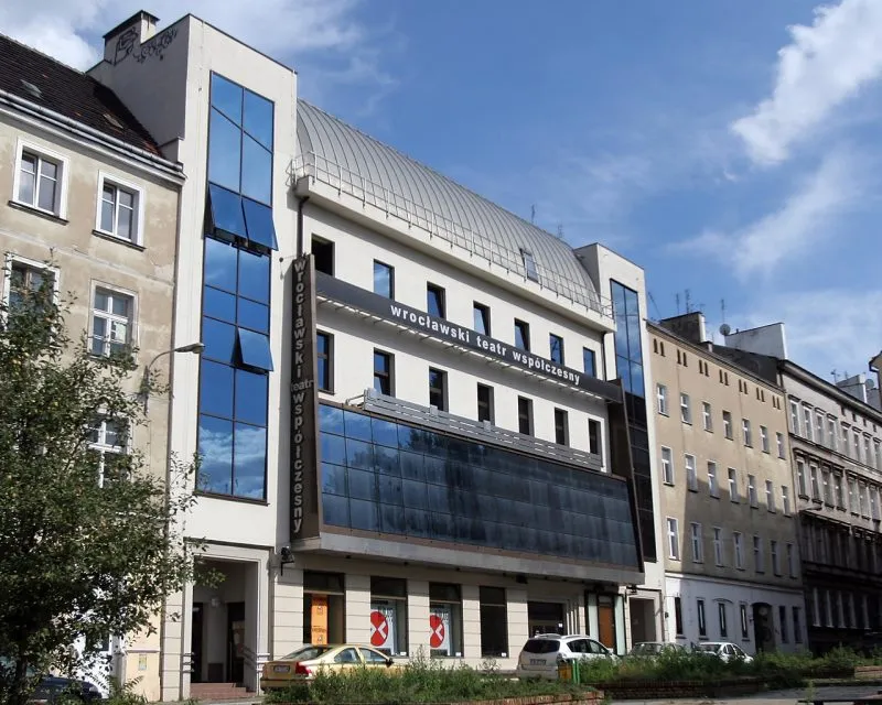 Nowoczesna fasada Wrocławskiego Teatru Współczesnego z ciemnym przeszklonym wejściem i pionowym napisem wypełnia pierzeję między starszymi kamienicami.
