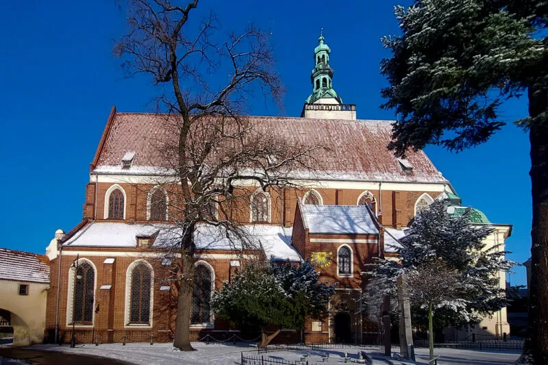 Zabytkowy gotycki kościół w Oleśnicy ma ceglane prezbiterium, wysoki czerwony dach i zieloną wieżę, otoczone ośnieżonym dziedzińcem.