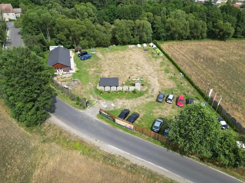 Niewielki kemping widziany z lotu ptaka obejmuje trawiastą polanę, domki, samochody i stanowiska wypoczynkowe przy drodze, na skraju gęstego lasu.