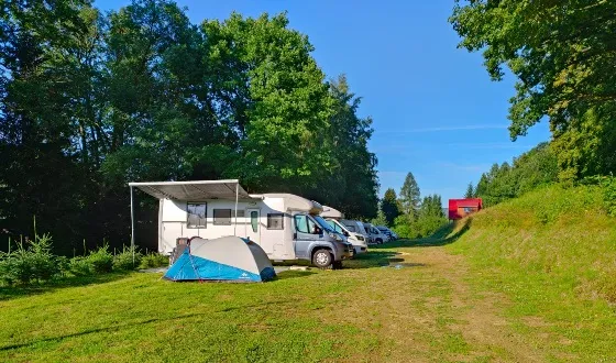 Kamper i przyczepy stoją na trawiastej parceli kempingowej przy zielonym wzgórzu, otoczone drzewami i otwartą przestrzenią do wypoczynku.