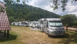 Kilka kamperów i przyczep kempingowych stoi na trawiastej parceli u podnóża zalesionego wzgórza, obok wiejskich zabudowań i ogrodzenia.