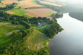 Widok z lotu ptaka ukazuje kemping nad dużym jeziorem, otoczony polami, wiejskimi zabudowaniami i mozaiką zielonych gruntów.