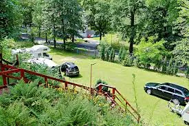 Niewielkie pole caravaningowe z samochodami i namiotami leży na zielonej polanie nad strumieniem, do której prowadzi czerwony drewniany mostek.