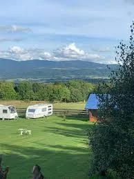 Zielone pole caravaningowe z przyczepami i niewielkim domkiem leży na wzgórzu, z którego rozciąga się szeroki widok na dolinę i odległe góry.