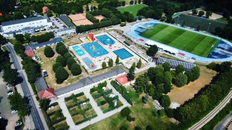 Widok z lotu ptaka na kompleks rekreacyjno-sportowy z basenami, boiskiem piłkarskim, kortami i geometrycznymi ogrodami pośród miejskiej zieleni.