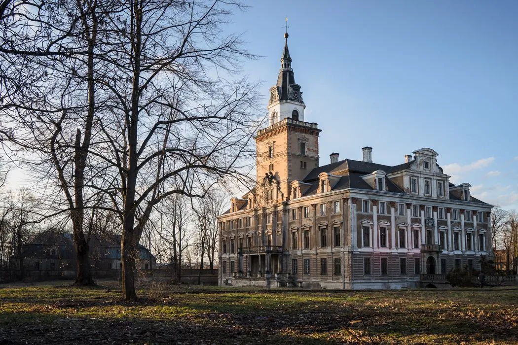 Rozległy Zamek Roztoka o jasnej, symetrycznej fasadzie i wysokiej wieży stoi wśród bezlistnych drzew, otoczony dawnym parkiem pod pogodnym niebem.