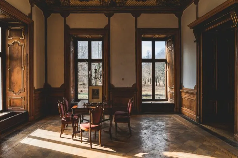 Reprezentacyjna sala Zamku Roztoka ma wysokie okna, ciemną drewnianą boazerię, ozdobne drzwi i niewielki stół z krzesłami ustawiony na zniszczonej podłodze.