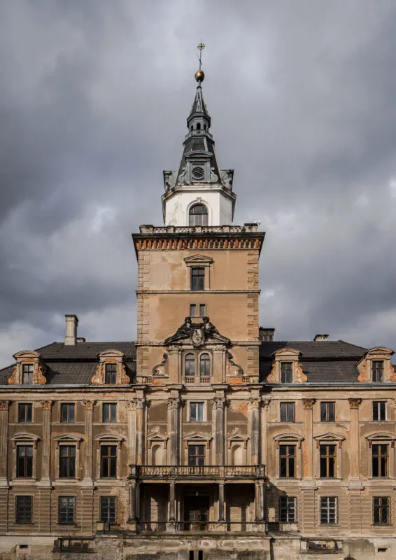 Reprezentacyjna fasada Zamku Roztoka z wieżą zegarową, mansardowym dachem i bogatym detalem architektonicznym wznosi się pod ciężkimi chmurami.