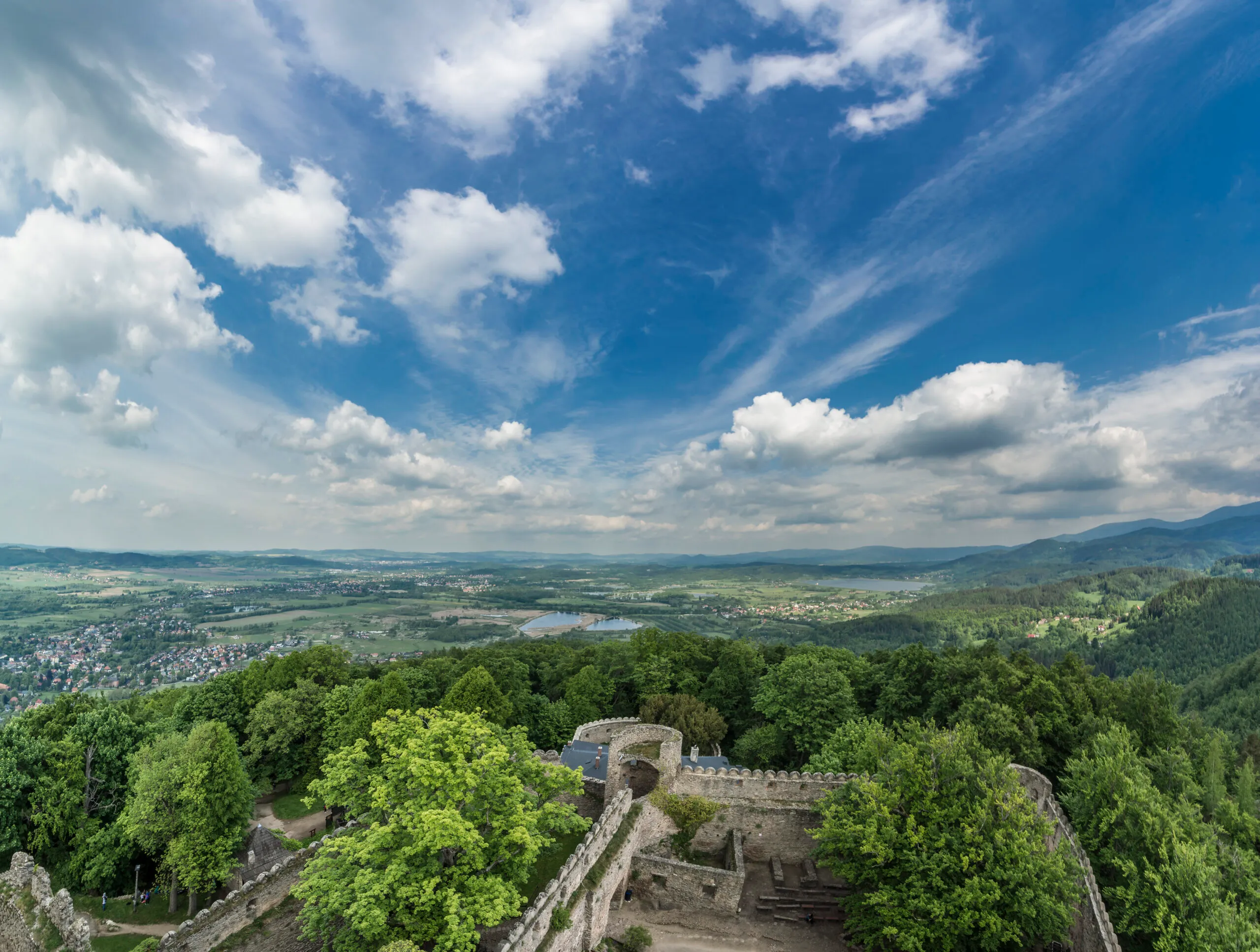 Widok znad murów kamiennego zamku otoczonego gęstym lasem. W tle ciekawa panorama najbliższej okolicy ze wzgórzami łąkami, stawem.