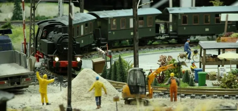 Makieta Kolejkowa przedstawia czarną lokomotywę parową z wagonami, miniaturowych robotników, maszyny budowlane i fragment kolejowego zaplecza.