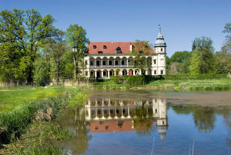 Na pierwszym planie znajduje się tafla jeziora, w której odbija się stojący nad jeziorem pałac. Pałac jest biały, ma czerwony dach oraz dwie wieże w narożnikach, a widoczna fasada jest bogata w arkady. Na trawniku, który jest po lewej stronie zdjęcia, widać kwitnące żółte kwiaty.